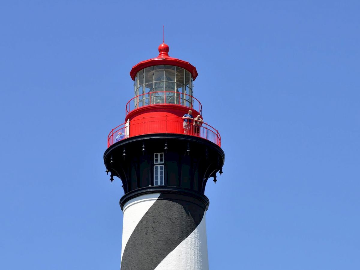 The image shows a lighthouse with black and white spiral stripes, topped with a red light, standing against a clear blue sky and surrounded by greenery.