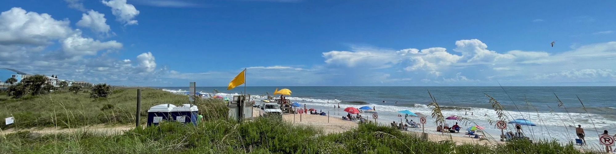 A beach scene with people, colorful umbrellas, green shrubs, yellow safety flag, and a partly cloudy blue sky.