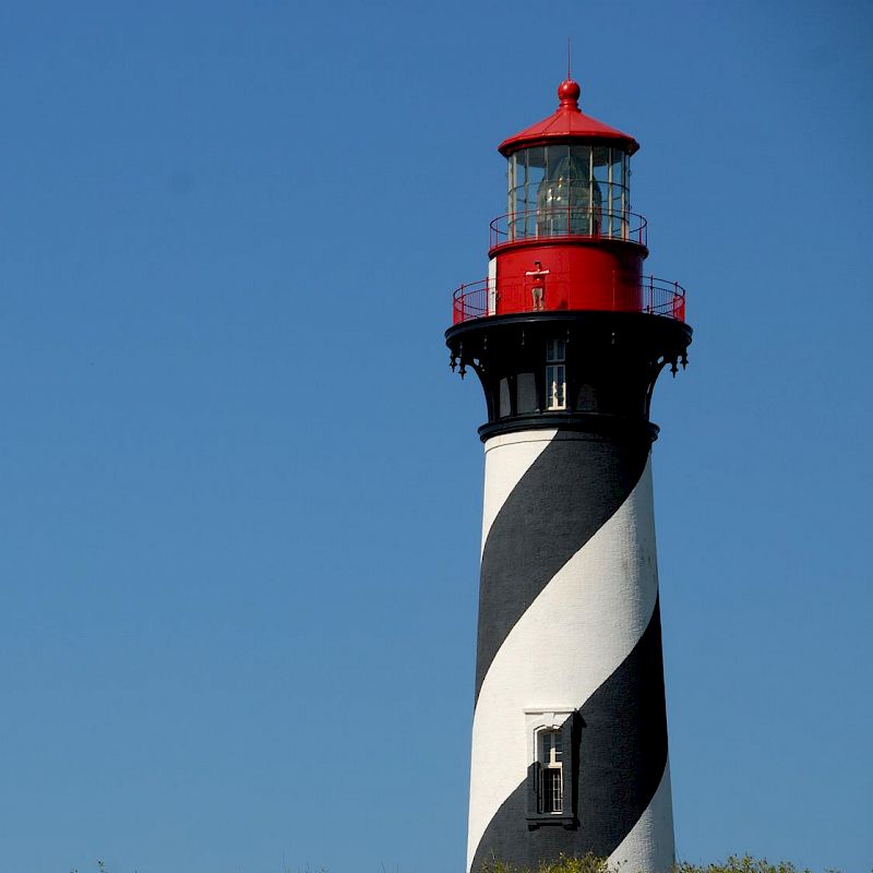 The image depicts a black and white striped lighthouse with a red top, set against a clear blue sky and surrounded by green trees.