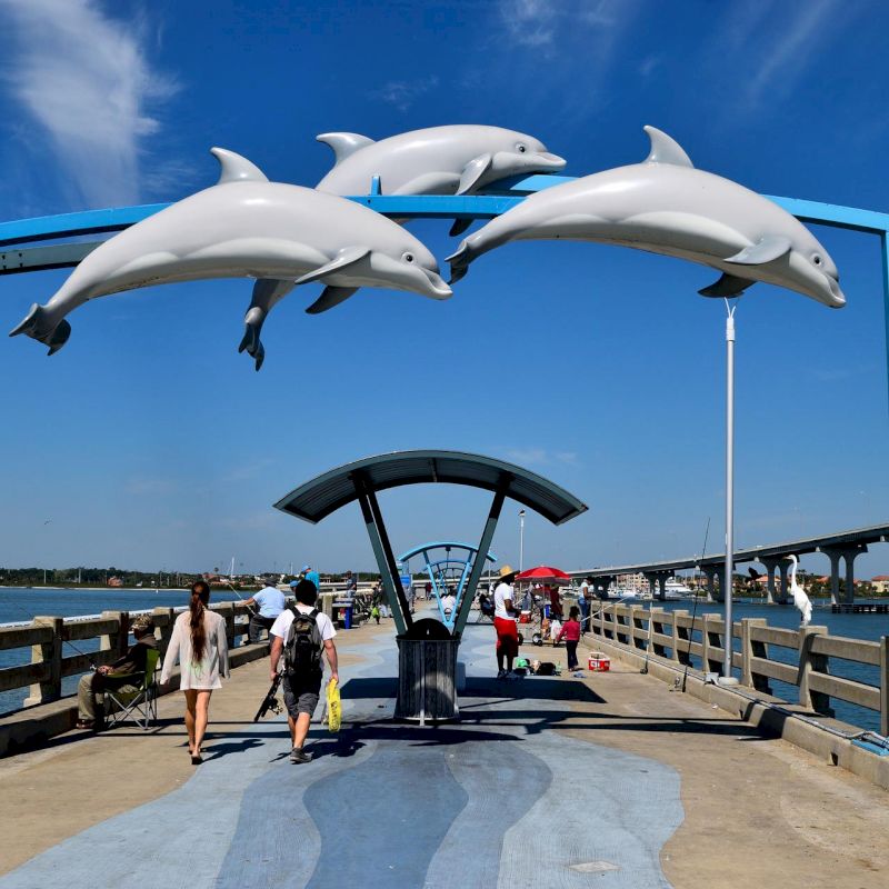 A pier with people walking under a decorative arch featuring three dolphin statues, with a body of water and bridge in the background, ending the sentence.