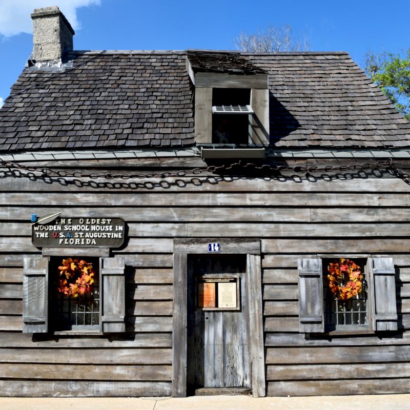 This image shows an old, historic wooden house with an American flag and autumn wreaths, identified as the oldest wood school house in Florida.