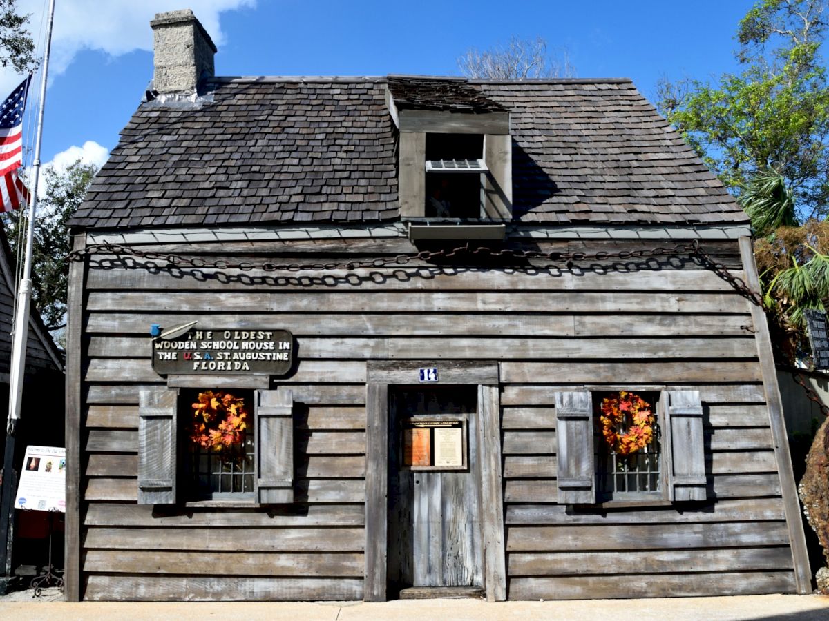 An old wooden schoolhouse with an American flag, decorated with wreaths, showcasing historical significance in a bright outdoor setting.