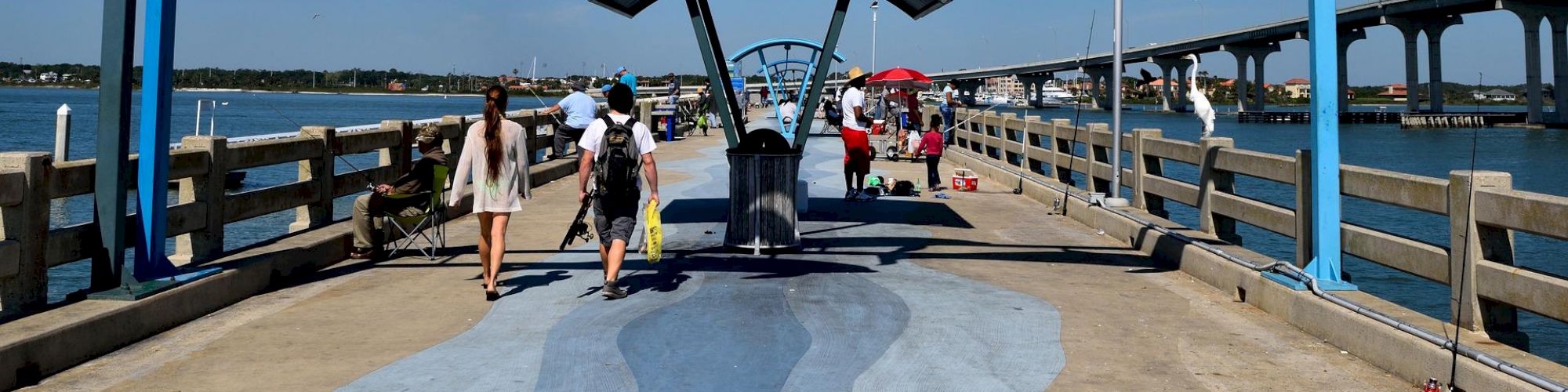 The image shows a walkway over water with a blue arch and three dolphin sculptures above, with people walking toward a covered seating area.