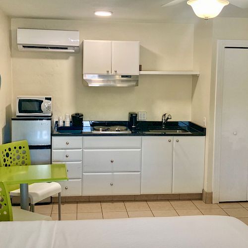 The image shows a small kitchen area with white cabinets, a sink, microwave, stovetop, and a green chair with a small table.