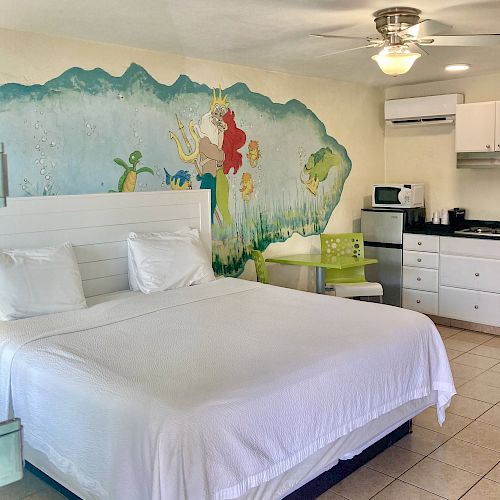 The image shows a hotel room with a large bed, ocean-themed mural on the wall, a ceiling fan, kitchenette, microwave, coffee maker, and a small dining area.