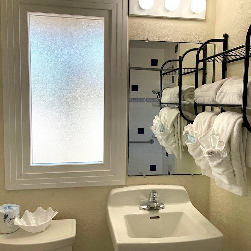 This image shows a bathroom with a window, sink, wall-mounted towel rack with rolled towels, a mirror, and a toilet with a tissue holder.