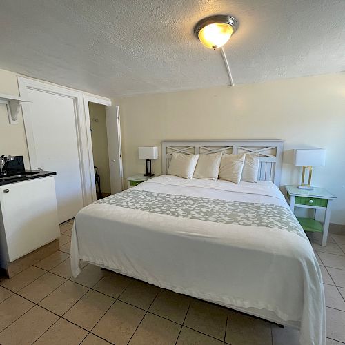 A room with a bed, nightstands, lamps, a mini fridge, a microwave, and a small kitchenette area, with a light fixture on the ceiling.