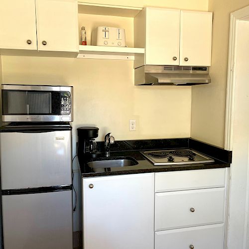 A compact kitchen with a mini-fridge, microwave, coffee maker, small sink, stove, overhead cabinets, and countertop, with a hallway leading to another room.