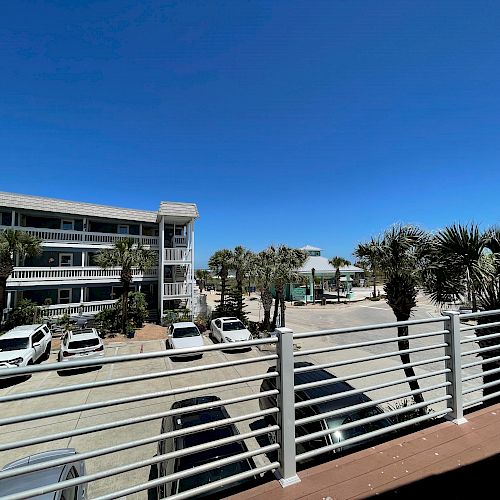 The image shows a coastal resort with parked cars, a multi-story hotel, palm trees, and a clear blue sky. A railing is in the foreground of the scene.