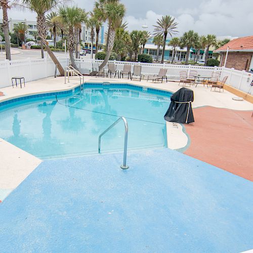 A small outdoor pool surrounded by a fence, deck chairs, and palm trees, with a grill covered in the background.