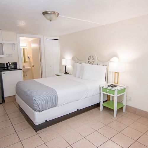 A hotel room with a bed, kitchenette (microwave, sink, coffee maker, mini-fridge), adjacent bathroom, bedside tables, and a green dresser.