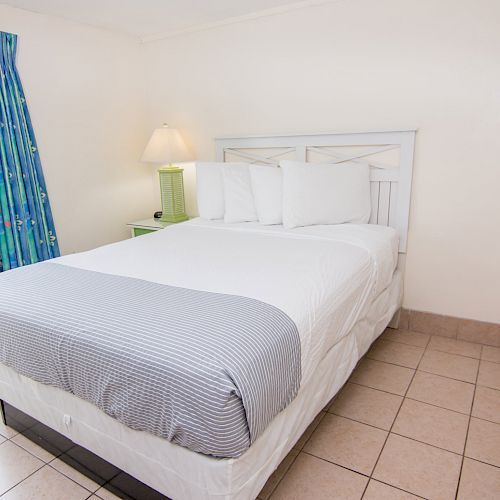 A small bedroom with a neatly made bed, white linens, a bedside lamp, colorful curtains, tile floor, and a picture on the wall.