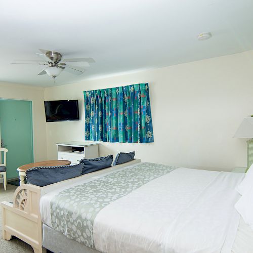 A brightly lit hotel room with a large bed, tropical-themed curtains, a wall-mounted TV, a ceiling fan, and a small table with chairs.