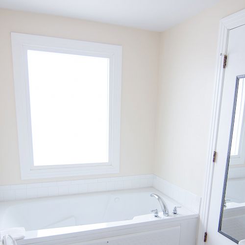 A bathroom with a large bathtub under a window, a door with a mirror, and light-colored walls is shown in the image.