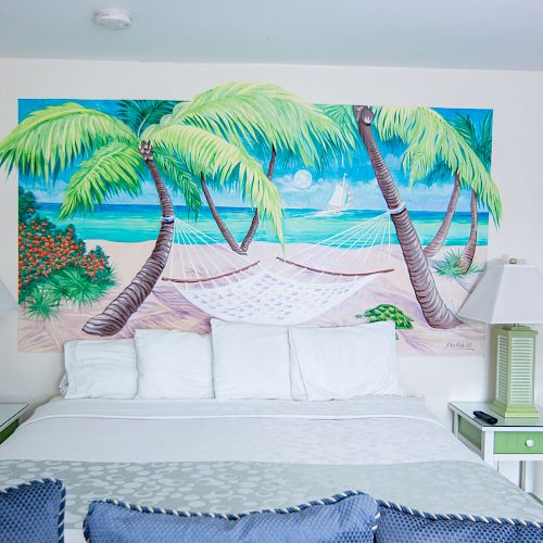 A bedroom with a beach-themed mural of palm trees and a hammock, featuring a bed with white linens and green bedside tables with matching lamps.