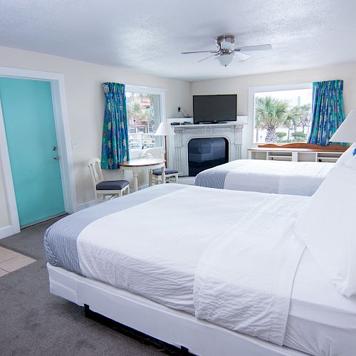 A cozy hotel room featuring two beds, white linens, a desk with a chair, TV, ceiling fan, blue curtains, and a turquoise door, with a window view.