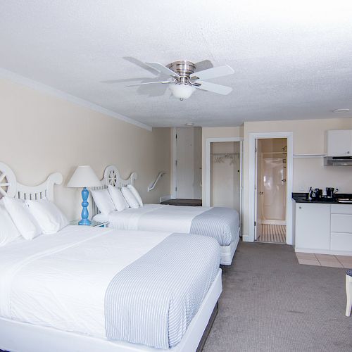 The image shows a clean, well-lit hotel room with two queen beds, a small kitchenette, a round table with chairs, and white furnishings throughout.