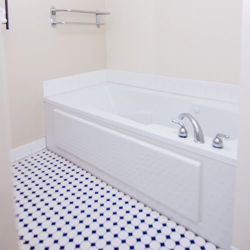 The image shows a bathroom with a rectangular bathtub, a white tiled front panel, and a polished bathroom floor with a blue and white pattern.