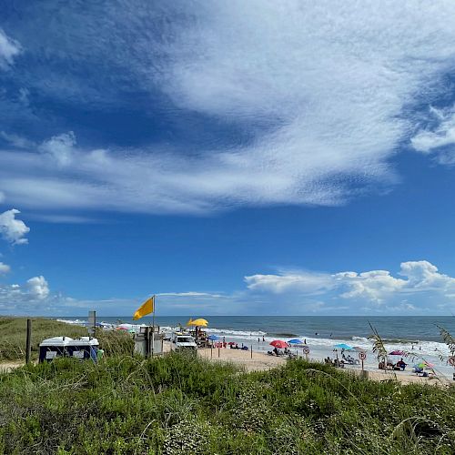 A beach scene with people under umbrellas, a yellow flag flying, grassy dunes in the foreground, and a partly cloudy sky above ending the sentence.