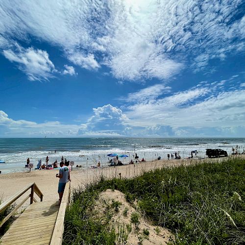 A beach scene with people enjoying the sand and ocean, some under umbrellas. A wooden walkway leads down to the beach from the greenery.