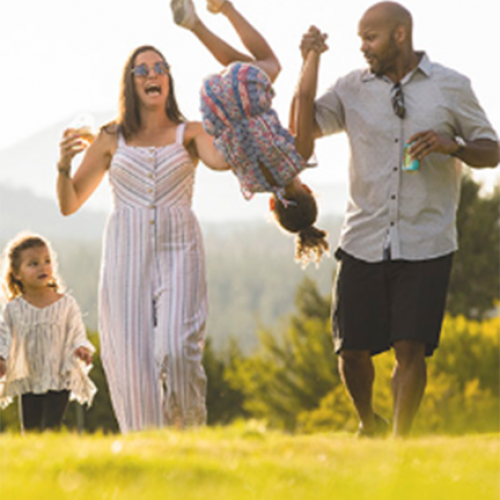A family of four enjoying a sunny day outdoors; parents holding hands with kids, one child swinging playfully in the air.