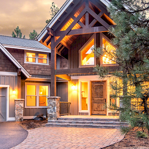 A large, picturesque home with glowing lights, surrounded by tall trees, featuring a three-car garage and a welcoming entrance.