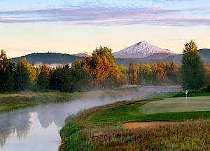 A serene landscape with a river, a golf course, trees, and a snow-capped mountain in the background under a partly cloudy sky at dawn.