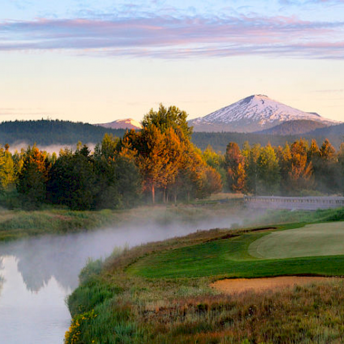 A serene landscape with a river, a golf course, trees, and a snow-capped mountain in the background under a partly cloudy sky at dawn.
