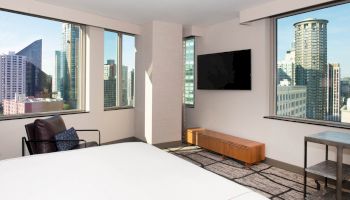 A modern hotel room with a bed, armchair, wall-mounted TV, and large windows showcasing a city skyline view.