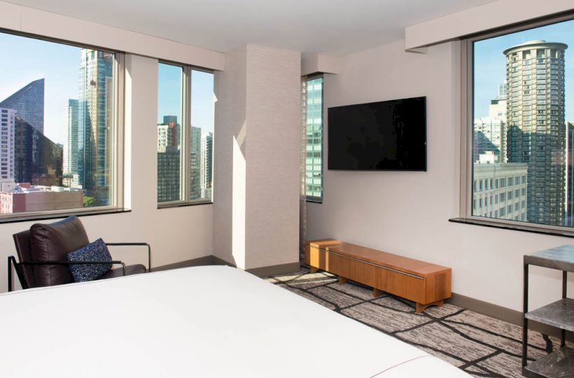 A modern hotel room with a bed, armchair, wall-mounted TV, and large windows showcasing a city skyline view.