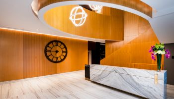 A modern interior with wood-paneled walls, a large clock, a winding staircase, a marble reception desk, and a flower arrangement.