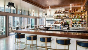 A modern bar featuring a well-stocked liquor shelf, elegant bar stools, and large windows overlooking a cityscape.