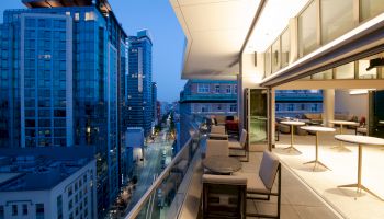 A modern outdoor terrace at twilight with tables, chairs, and cityscape views including tall buildings and a street below, ending the sentence.