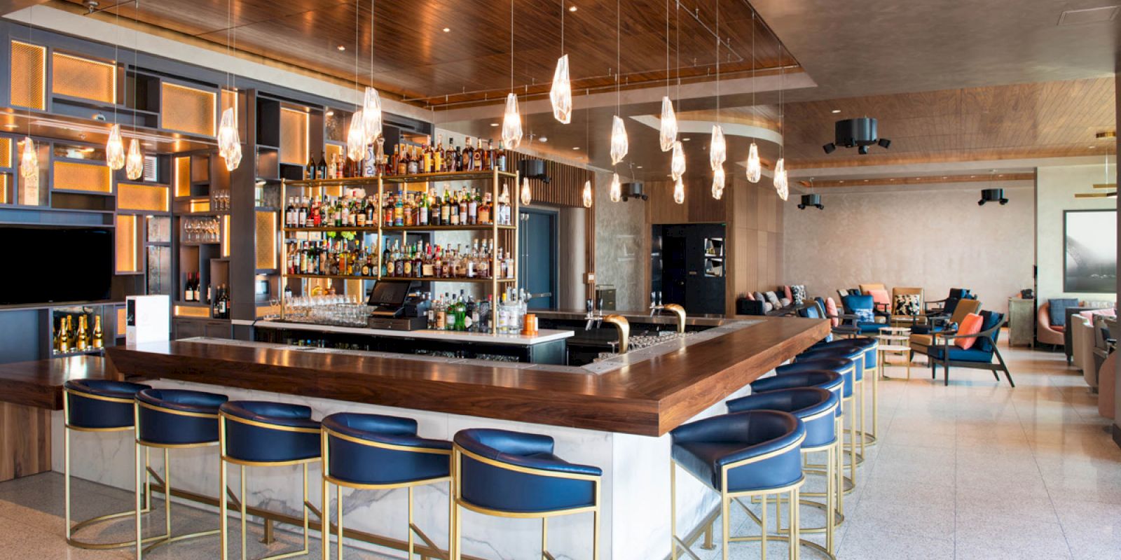 This image shows a modern bar with a well-stocked back shelf, blue bar stools, decorative lighting, and a cozy seating area in the background.