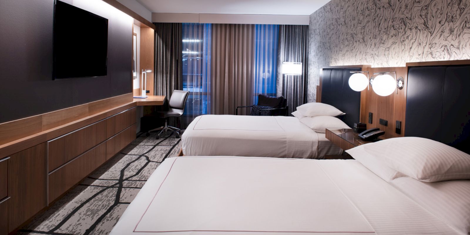 A modern hotel room with two neatly made beds, a flat-screen TV, a desk with a chair, and contemporary decor featuring patterned walls and curtains.