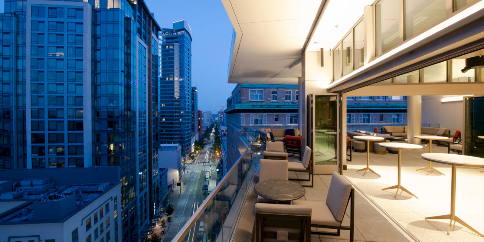 A modern urban balcony with stylish furniture overlooks a city street lined with tall buildings, illuminated by streetlights at dusk.