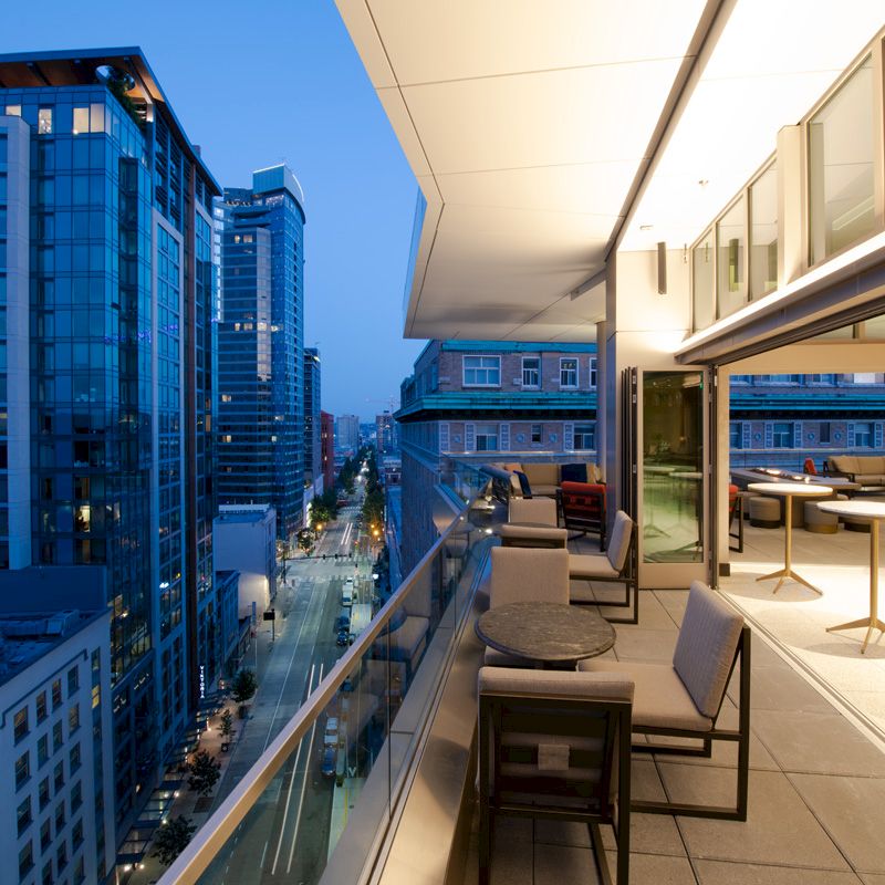 This image shows a modern cityscape view from a stylish balcony at dusk, featuring outdoor seating and tables with high-rise buildings in the background.