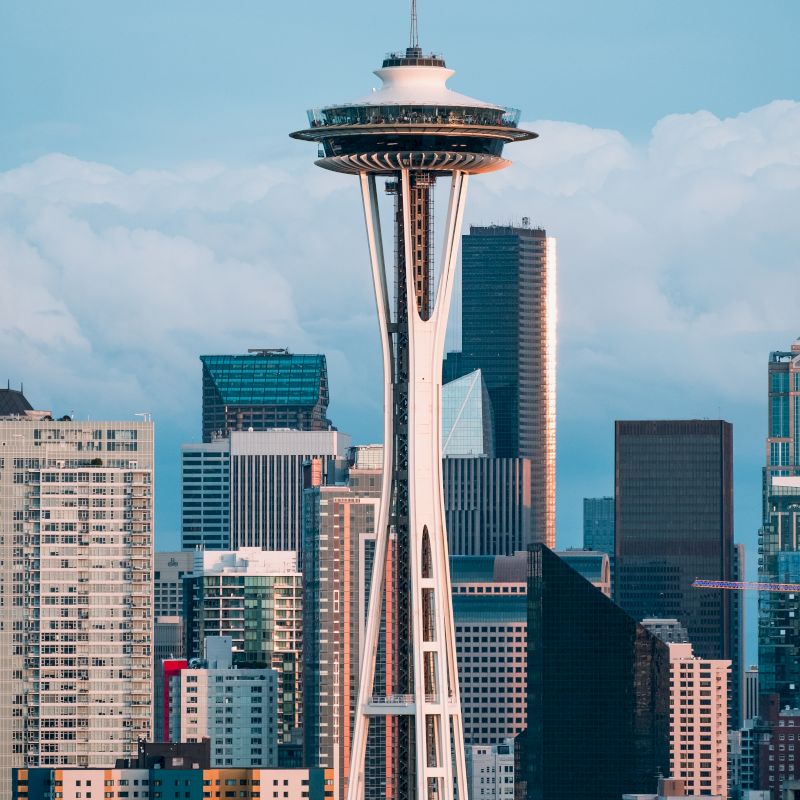The image shows the Space Needle tower in Seattle, Washington, surrounded by various buildings and skyscrapers under a cloudy sky.