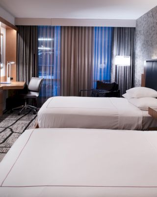 A modern hotel room with two beds, a desk, a chair, a large TV, and stylish decor featuring patterned wallpaper and curtains, is shown in this image.