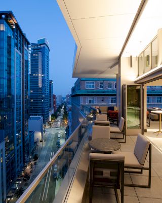 A modern cityscape at dusk with high-rise buildings and a balcony featuring outdoor seating and tables overlooking a street.