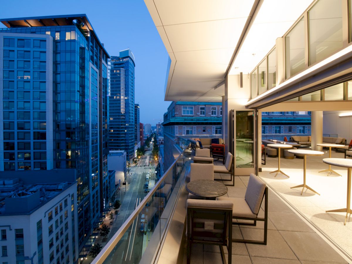 A modern, open balcony at dusk with city views, featuring elegant furniture and glass railings, connected to an indoor area with tables and chairs.