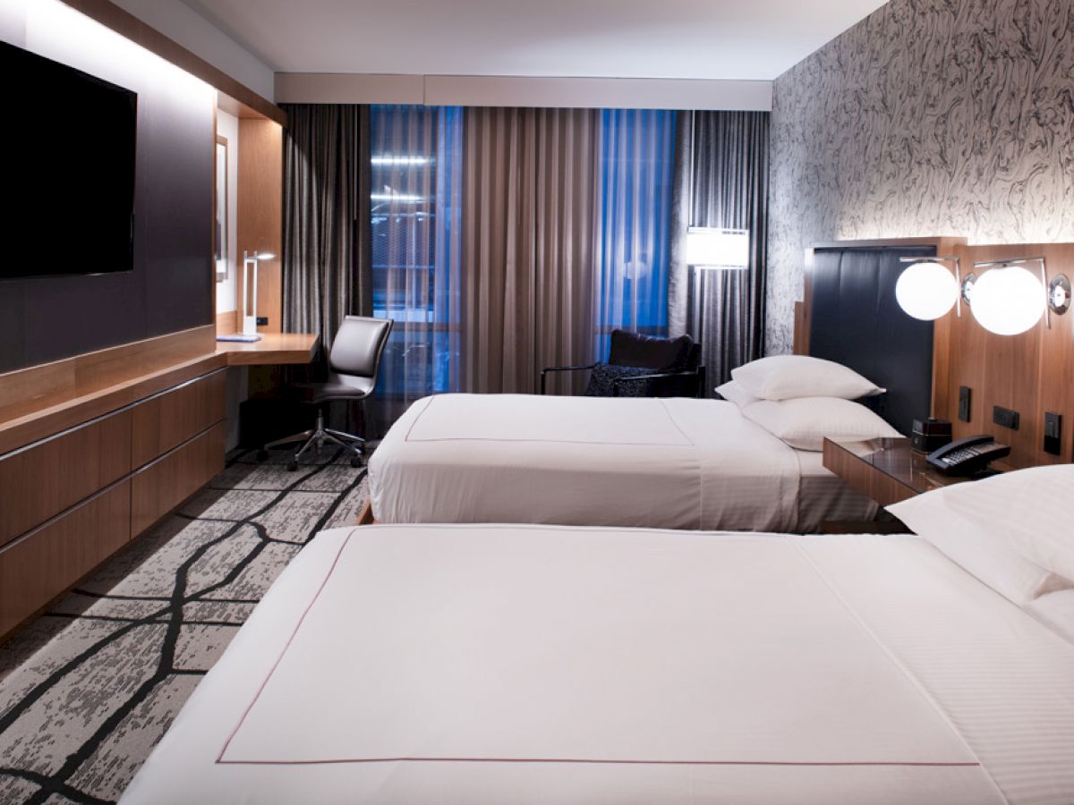 This image shows a hotel room with two beds, desk, chair, large TV, and modern lighting and decor, including patterned wallpaper and floor.
