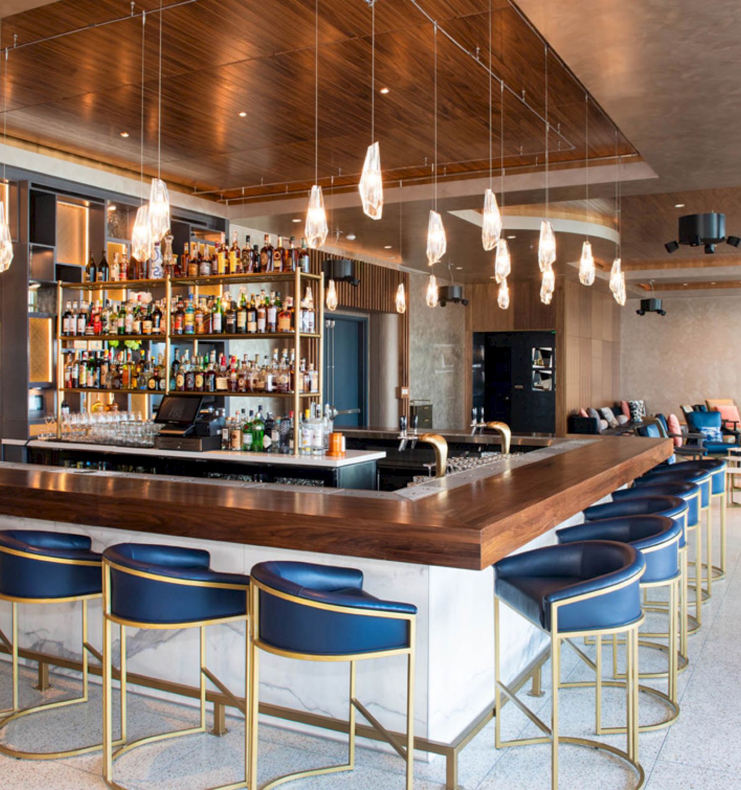A modern bar with a wooden counter, blue barstools, a fully stocked backbar, pendant lights above, and a seating area in the background.
