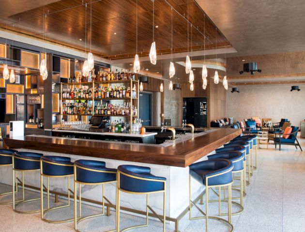 A modern bar with high stools, a hanging light fixture, and a well-stocked shelf of bottles; lounge seating is visible in the background.