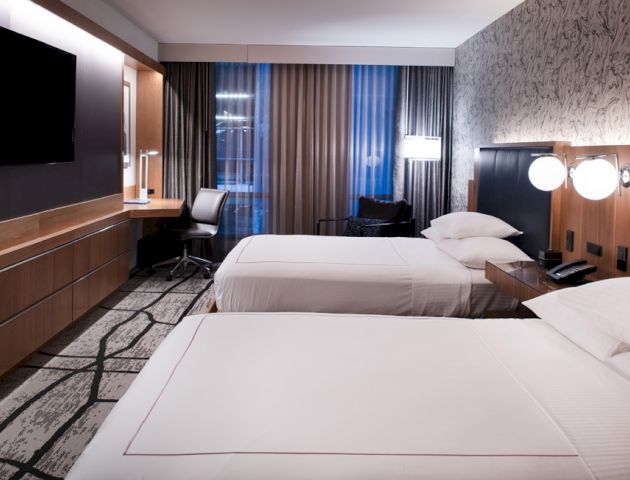A modern hotel room with two beds, a flat-screen TV, a desk, lamps, and a large window with curtains.