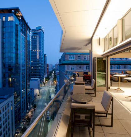 A modern balcony with seating and tables overlooks a cityscape filled with tall buildings at dusk.