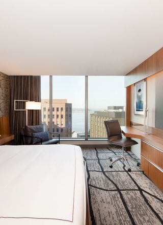 A modern hotel room features a large bed, a desk, a chair, large windows with a city view, and a flat-screen TV mounted on a wooden wall unit.