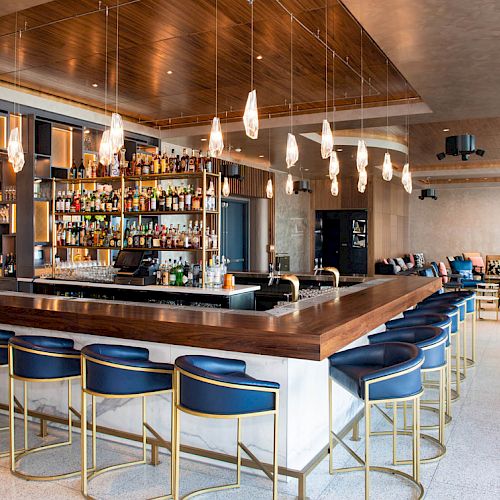This image shows a modern bar area with a large counter, cushioned stools, various bottles of alcohol on display, and stylish lighting fixtures.
