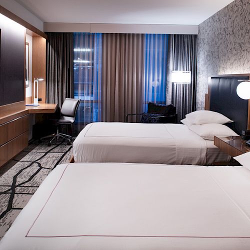 The image depicts a modern hotel room with two neatly made beds, a flat-screen TV, a desk, and chairs, lit with stylish lamps.