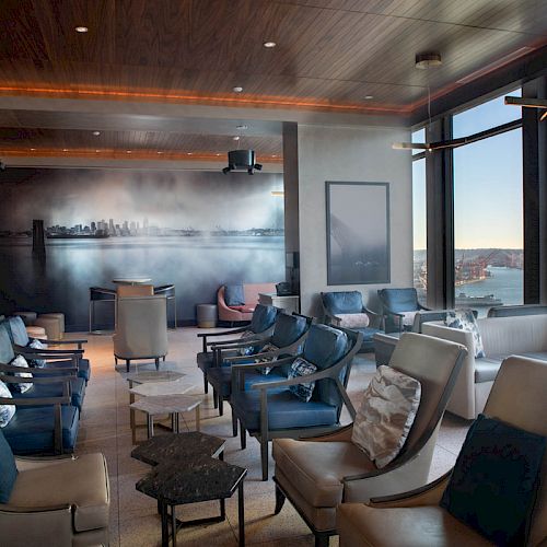A modern, comfortable lounge area with large windows offering a scenic view of a waterfront and a ferris wheel, featuring ample seating.